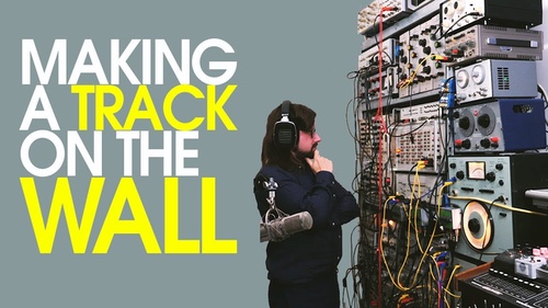 Person in black with headphones stands thoughtfully before a wall of audio equipment and tangled wires. Text reads “Making a Track on the Wall.”